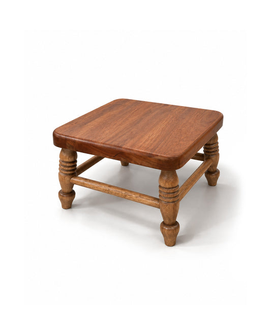 A small square wooden stool with a medium brown finish, featuring turned legs and a lower support rail, photographed on a white background.